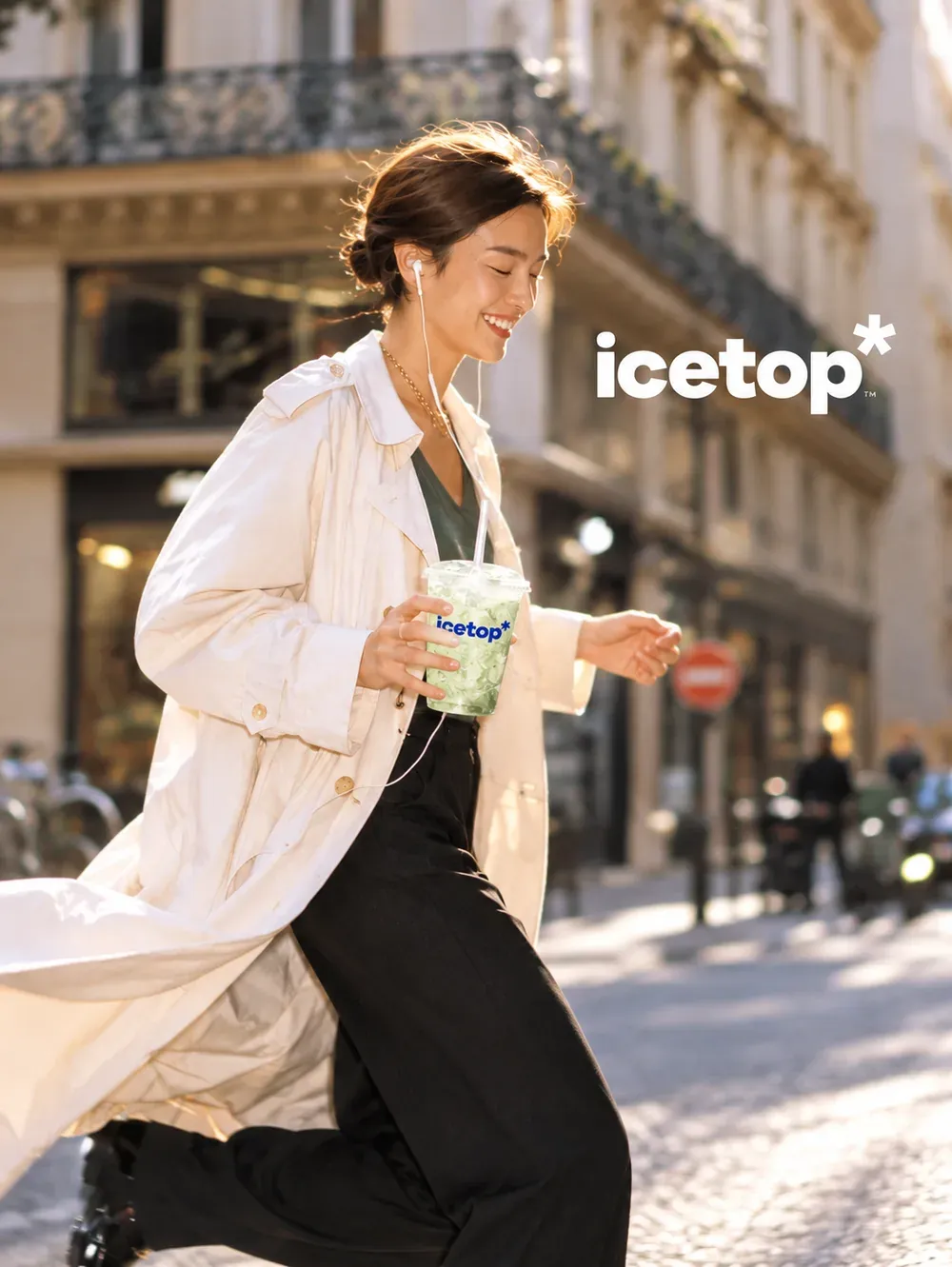 A woman in a cream trench coat walking down a sunny street, smiling, holding an icetop cup with a green iced drink